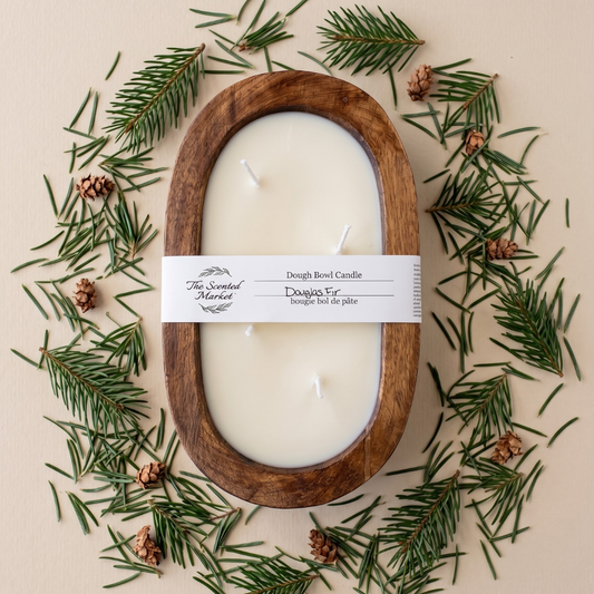 A wooden oval candle holder filled with white wax, wrapped with a white paper label reading 'The Sacred Melt' and 'Dough Bread Candle'. The candle is arranged in a flat lay on a beige background, surrounded by pine needles and small pinecones.