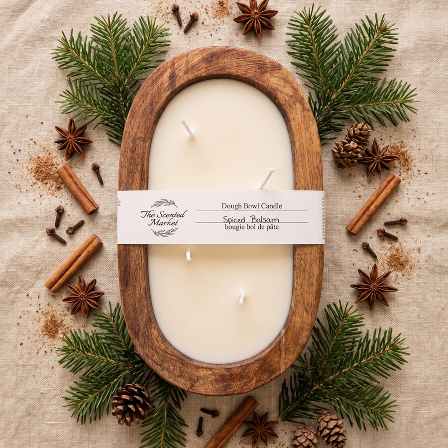 An oval wooden candle holder filled with cream-colored wax, wrapped with a white paper band labeled 'The Scented Market Dough Bowl Candle.' The candle is arranged in a styled flat lay on a beige fabric background, surrounded by green pine branches, star anise, cinnamon sticks, cloves, and pinecones.